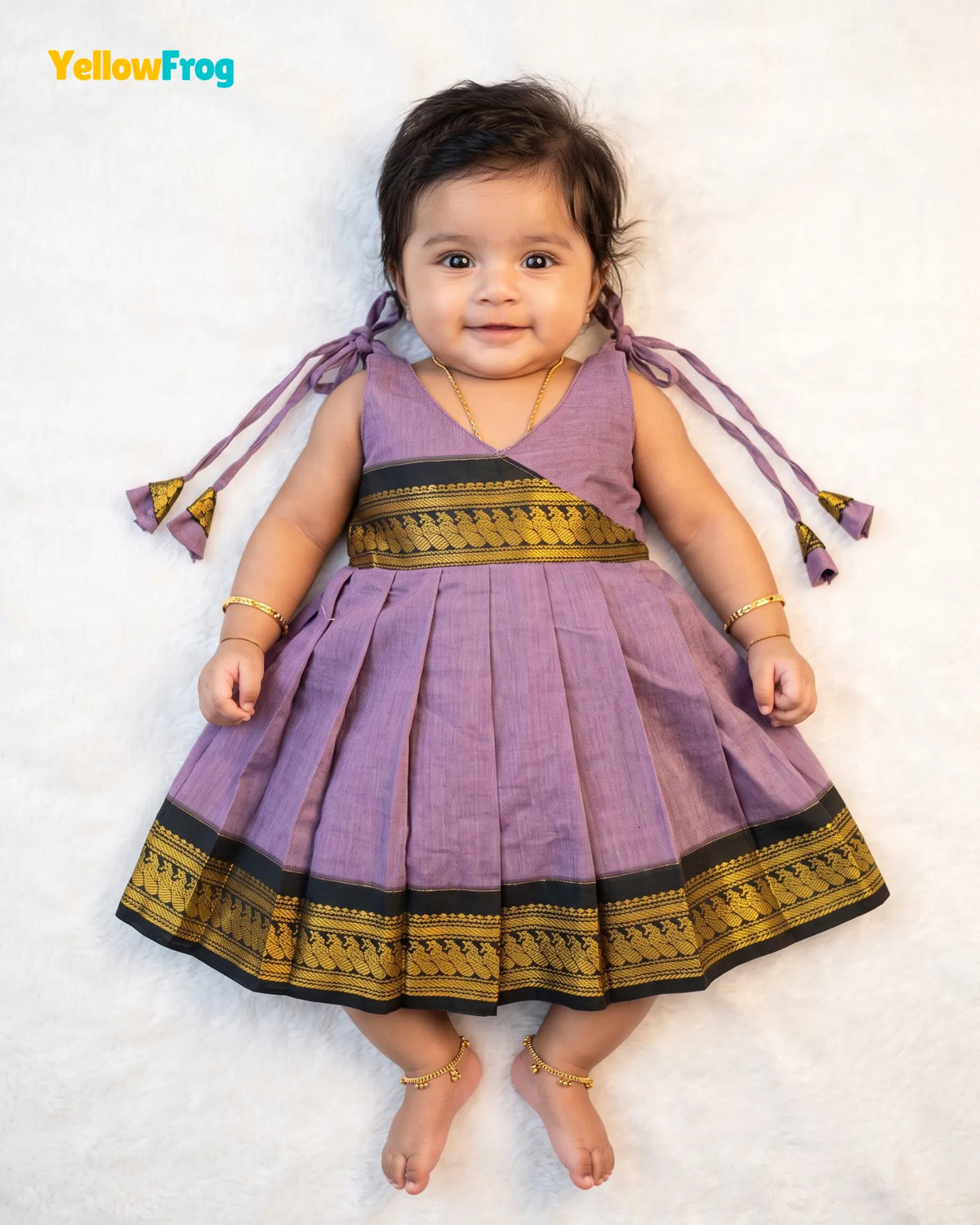A toddler girl lying on a white textured blanket, wearing a sleeveless lavender purple traditional dress with a pleated skirt and gold and black borders. The dress has purple ribbon ties on the shoulders.
