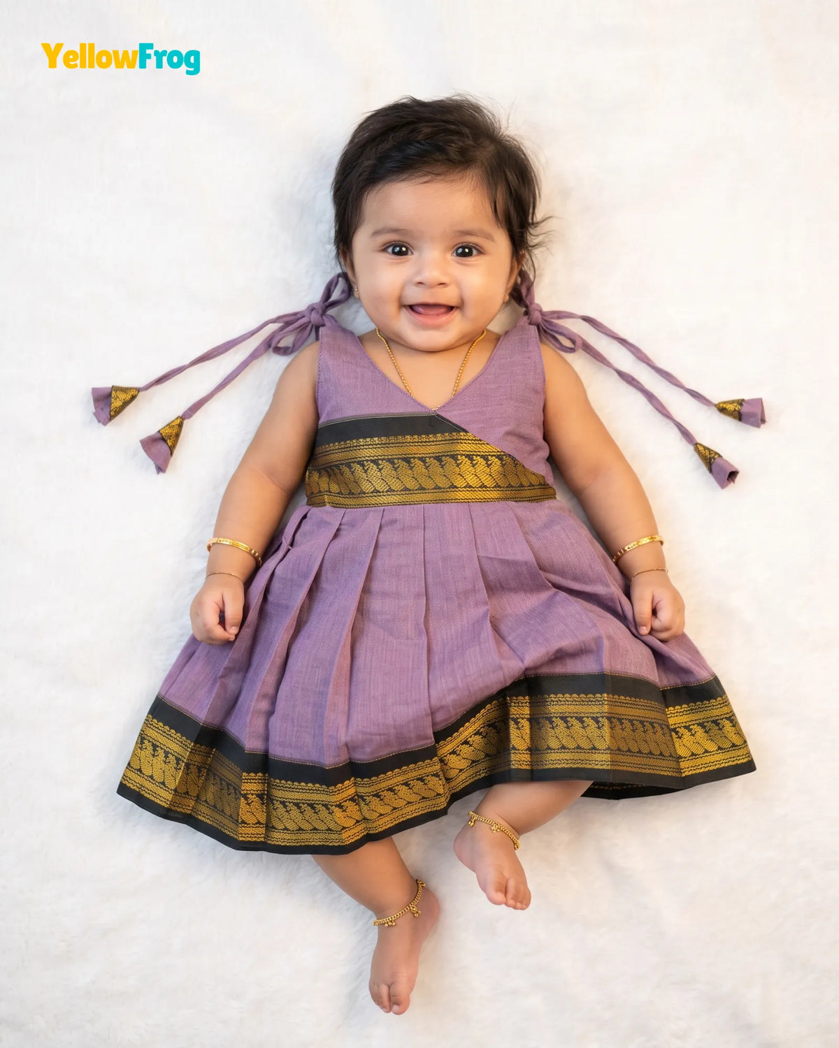 A baby girl lying on a white textured blanket, wearing a sleeveless lavender purple dress with gold and black patterned borders and tie details on the shoulders. The YellowFrog logo is visible in the top left corner.