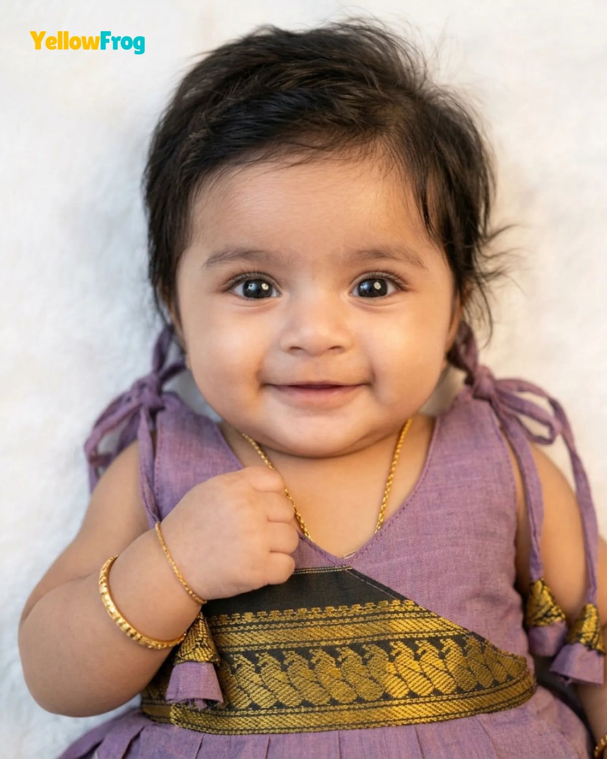 A smiling toddler with dark hair wearing a lavender purple traditional outfit with a gold patterned sash, a gold necklace, and a gold bracelet, set against a soft white textured background.