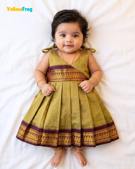 A baby girl lying on a white sheet wearing a yellow-green pleated traditional Indian dress with purple and gold borders. The brand name 'YellowFrog' is visible in the top left corner.