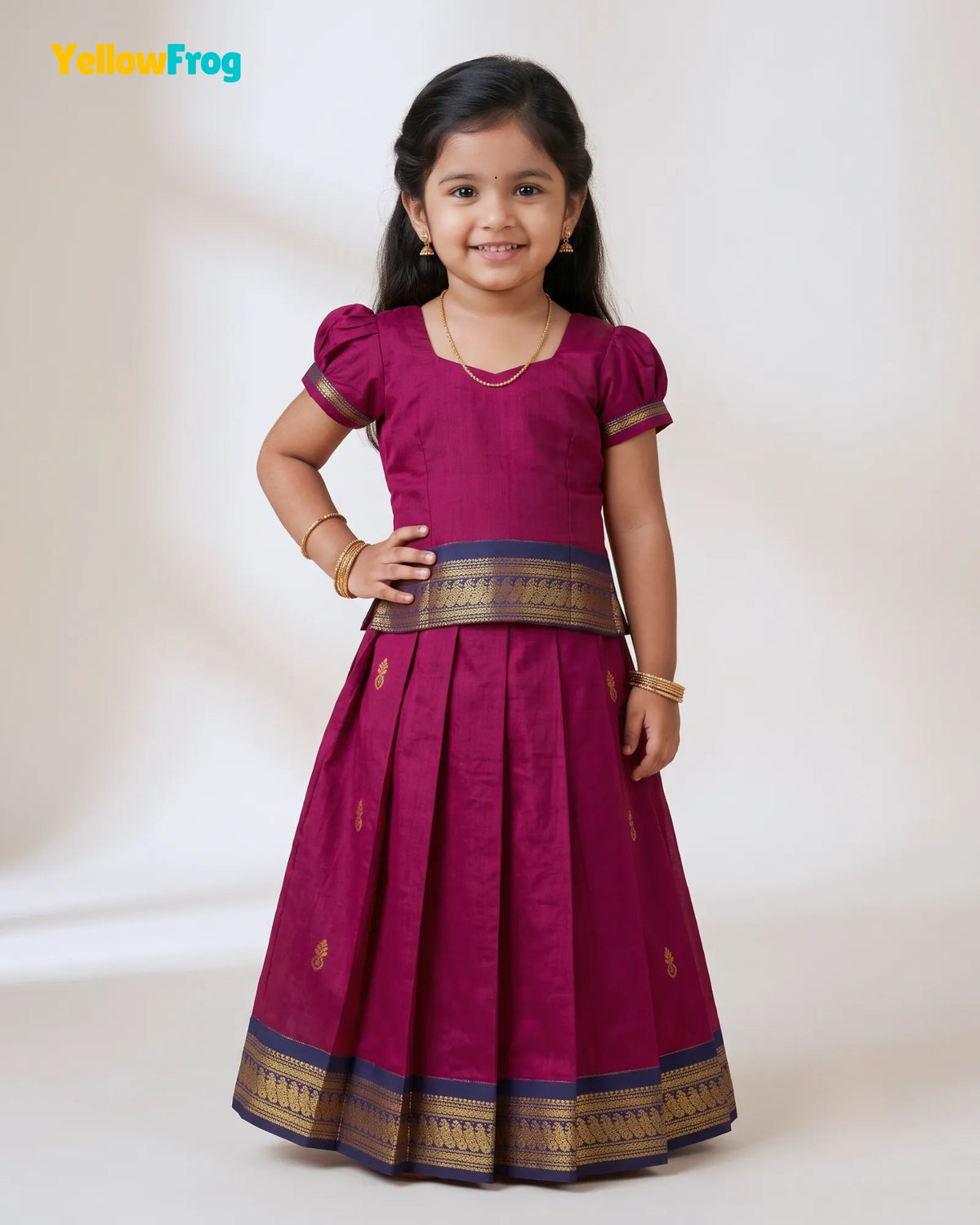 A young girl modeling a magenta traditional Indian dress with gold and navy borders, standing against a plain off-white studio background. The YellowFrog logo is visible in the top left corner.