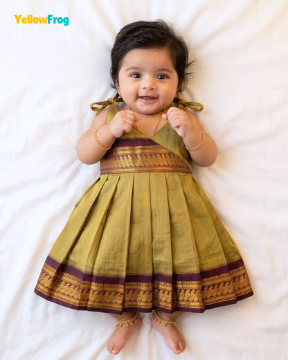 A baby girl lying on a white sheet wearing a yellow-green pleated traditional Indian dress with gold and purple borders. The brand name 'YellowFrog' is visible in the top left corner.