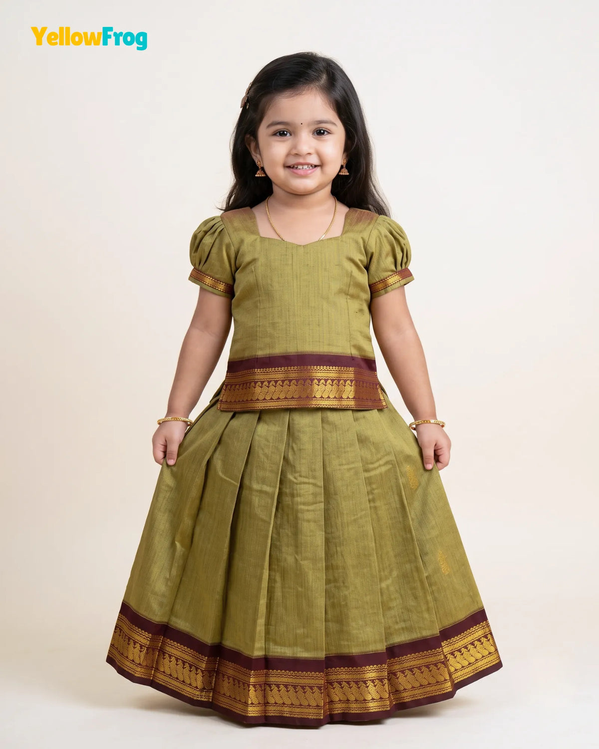 A young girl modeling a traditional Indian dress, featuring a muted olive green body with brown and gold zari borders, set against a plain light beige background. The YellowFrog logo is visible in the top left corner.
