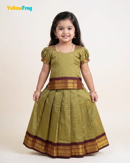 A young girl modeling a traditional Indian dress, featuring a muted olive green body with brown and gold zari borders, set against a plain light beige background. The YellowFrog logo is visible in the top left corner.