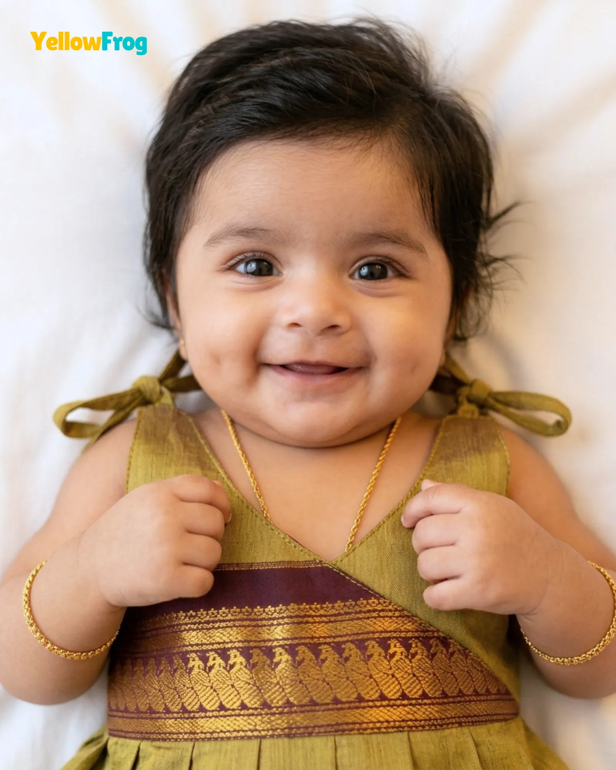 A smiling baby lying down wearing a traditional Indian dress in olive green and gold with a maroon border, along with gold jewelry. The YellowFrog brand logo is visible in the top left corner.