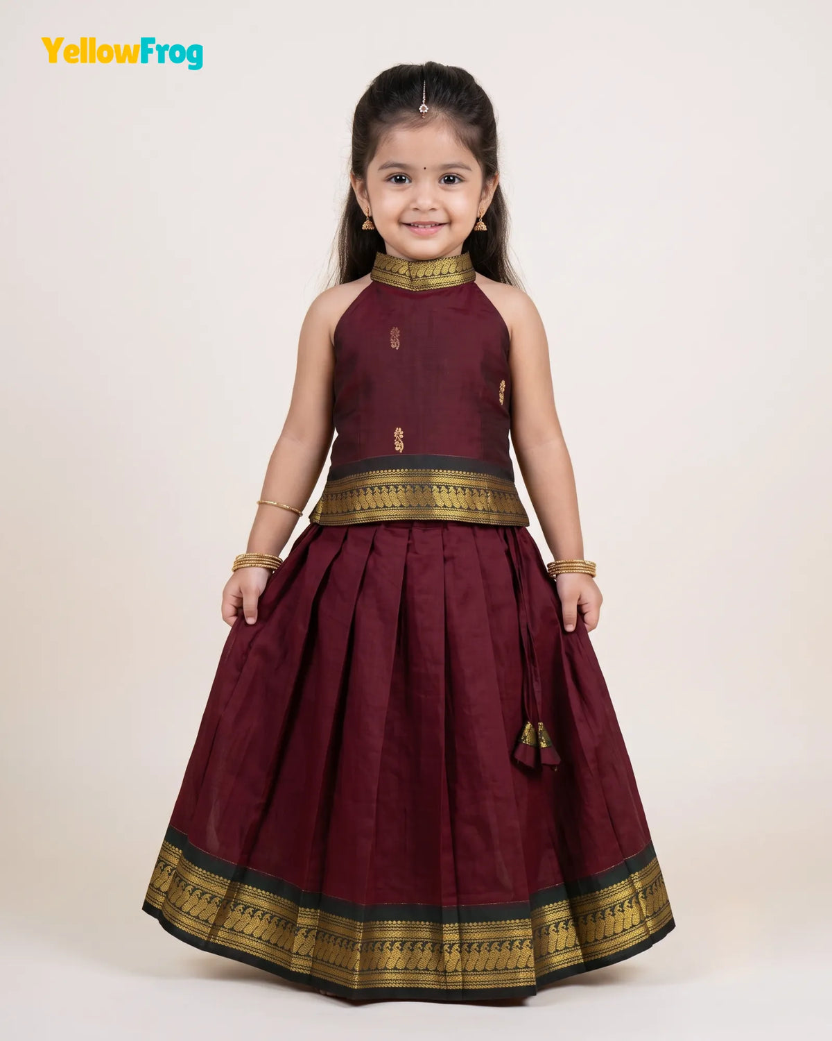 A young girl modeling a traditional Indian outfit consisting of a sleeveless maroon top and a matching pleated skirt with gold and black borders, set against a plain light beige background.