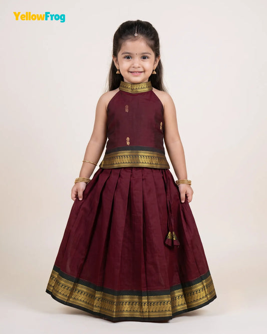 A young girl modeling a traditional Indian outfit consisting of a sleeveless maroon top and a matching pleated skirt with gold and black borders, set against a plain light beige background.