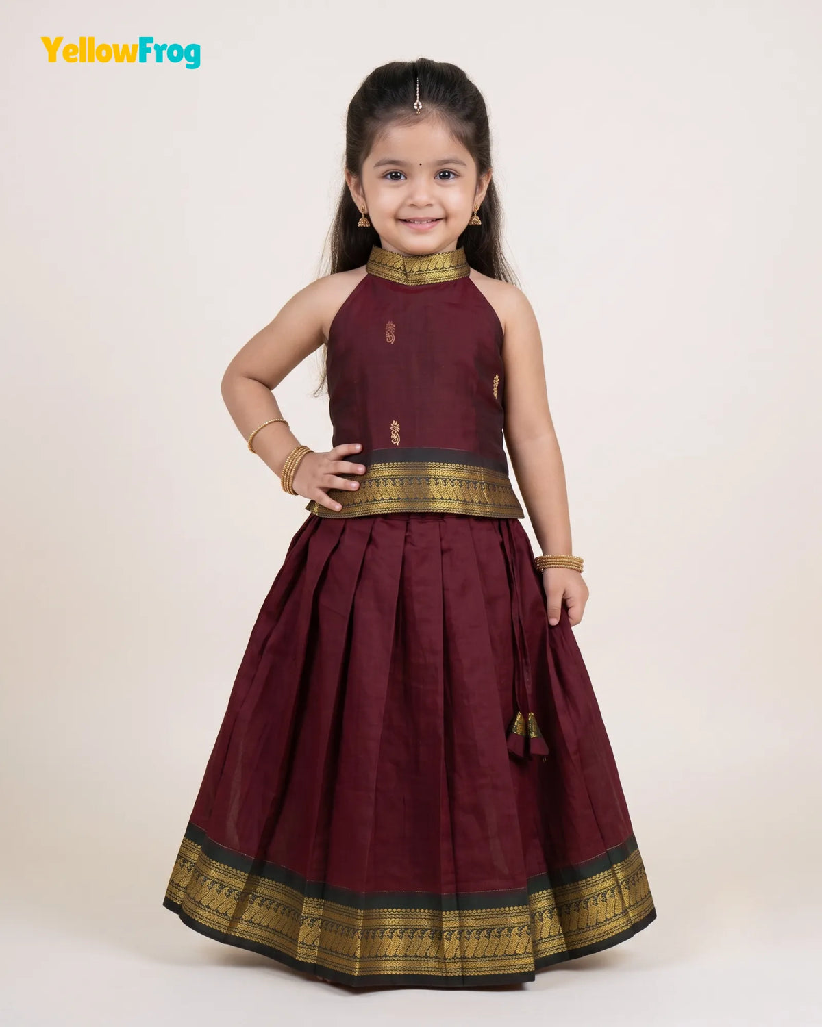A young girl modeling a traditional Indian outfit consisting of a sleeveless maroon lehenga skirt with gold zari borders and a matching maroon and gold choli top, set against a plain cream background.