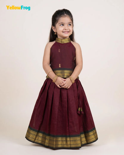 A young girl modeling a sleeveless maroon traditional dress with gold and black zari borders, standing against a plain cream background. The YellowFrog logo is visible in the top left corner.