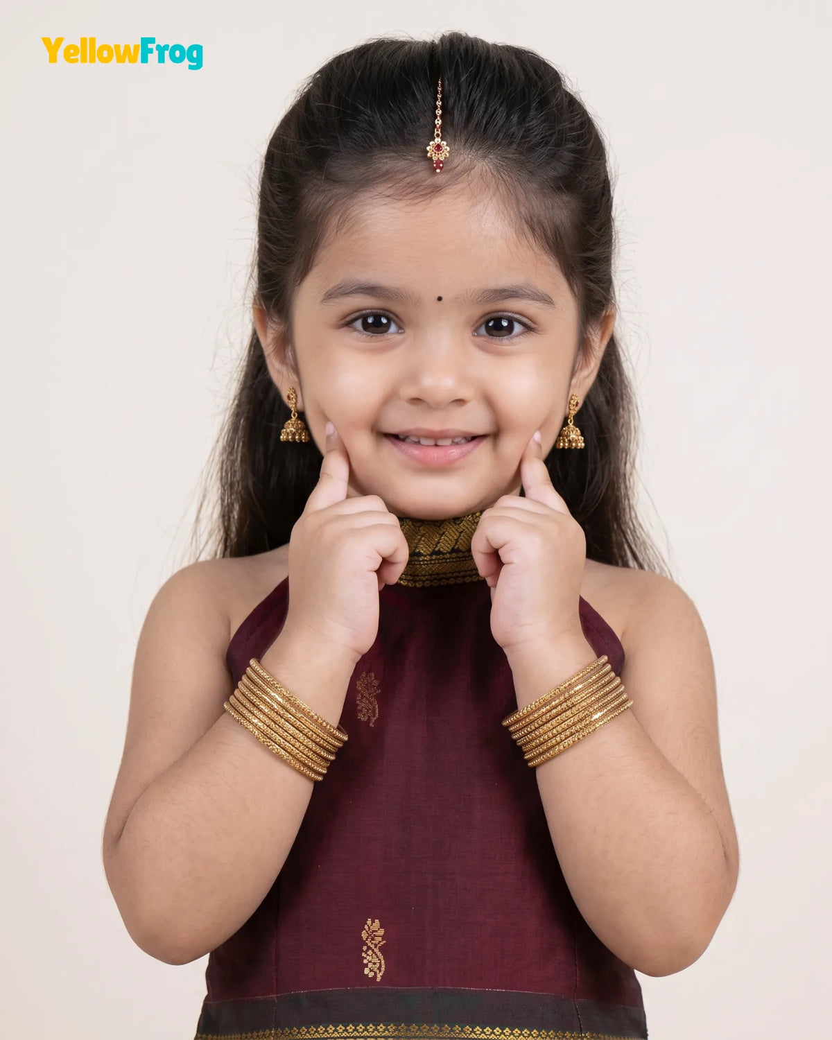 A young girl with dark hair styled in a bun, wearing a maroon traditional sleeveless top with gold trim and matching gold jewelry, posing with her hands on her cheeks against a light beige background. The YellowFrog logo is visible in the top left corner.
