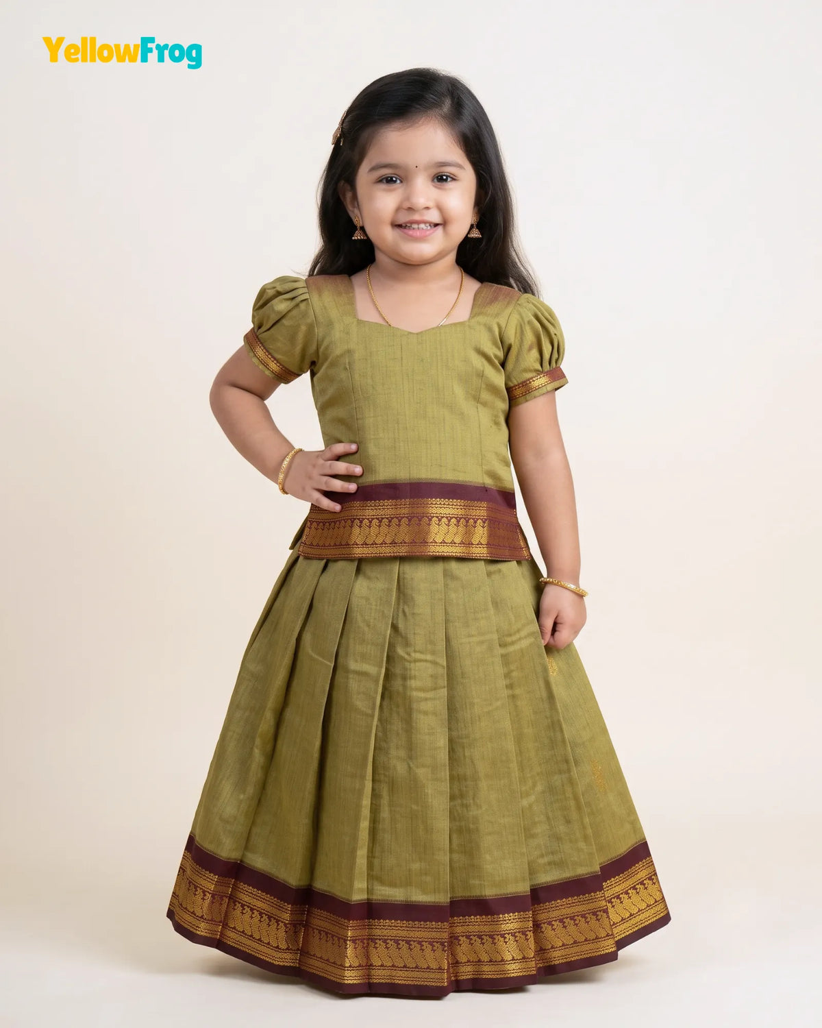 A young girl modeling a traditional Indian dress consisting of an olive green kurta with gold and maroon borders, paired with a matching pleated skirt. The image is set against a plain light beige background with a 'YellowFrog' logo in the top left corner.