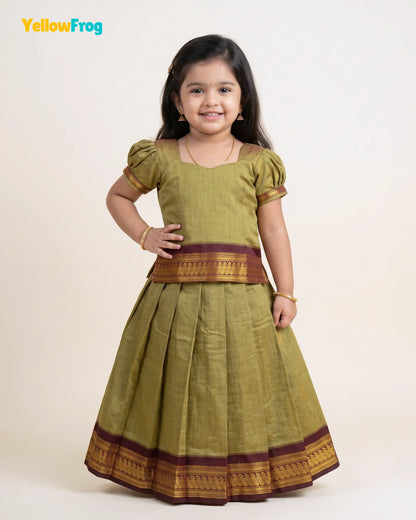 A young girl modeling a traditional Indian dress consisting of an olive green kurta with gold and maroon borders, paired with a matching pleated skirt. The image is set against a plain light beige background with a 'YellowFrog' logo in the top left corner.