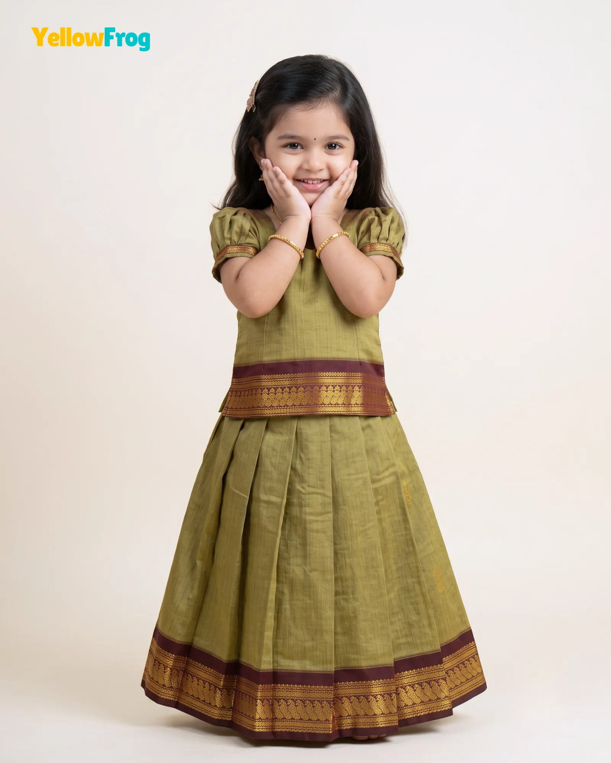 A young girl modeling a traditional Indian dress consisting of an olive green kurta with gold and maroon borders, paired with a matching pleated skirt. The YellowFrog brand logo is visible in the top left corner.