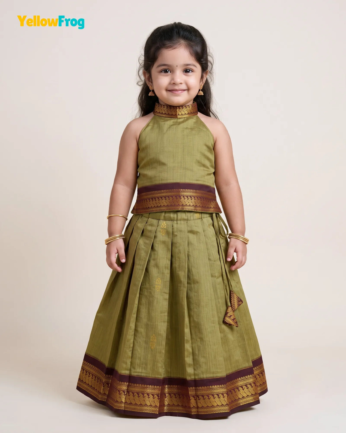 A young girl modeling a sleeveless olive green traditional dress with gold and maroon borders, standing against a plain beige studio background. The YellowFrog logo is visible in the top left corner.