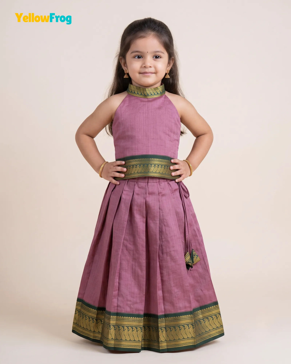 A young girl modeling a sleeveless dusty purple traditional dress with green and gold zari borders, standing against a solid light beige background. The YellowFrog brand logo is visible in the top left corner.