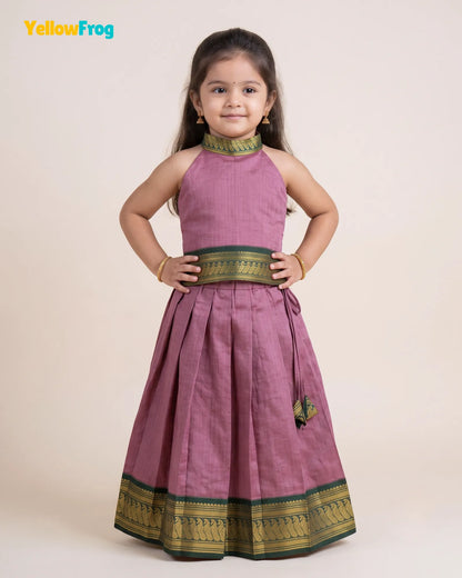 A young girl modeling a sleeveless dusty purple traditional dress with green and gold zari borders, standing against a solid light beige background. The YellowFrog brand logo is visible in the top left corner.