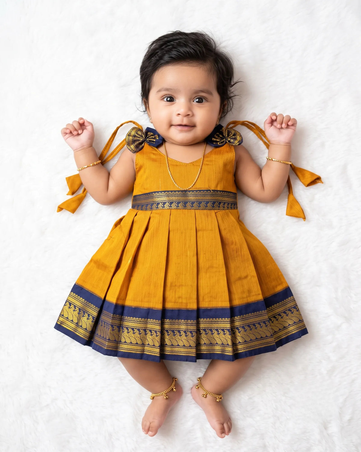A baby girl lying on a white textured blanket, wearing a mustard yellow and navy blue traditional Indian dress with gold accents.