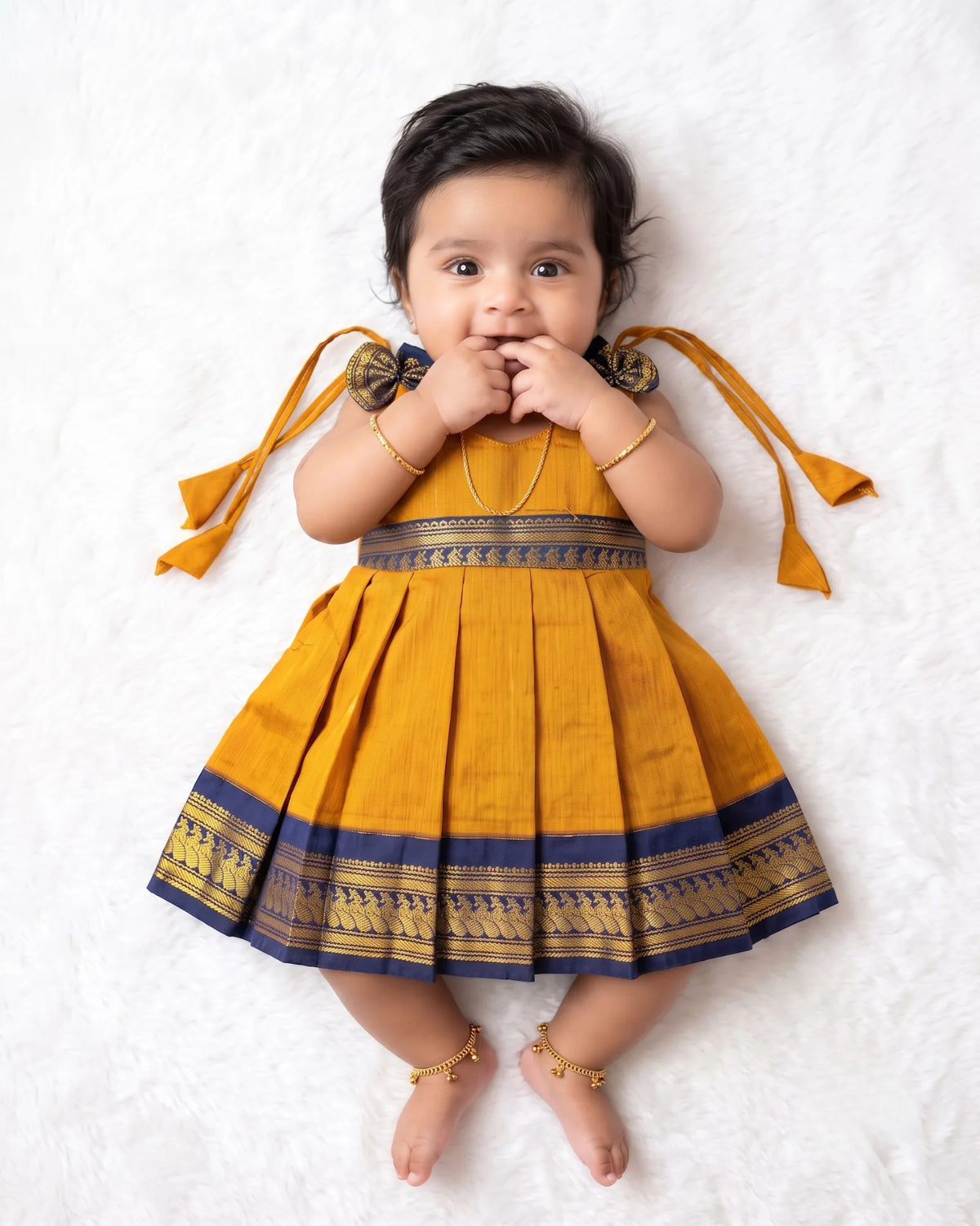 A baby girl lying on a white textured blanket, wearing a mustard yellow pleated traditional dress with navy blue and gold borders and tie details on the shoulders.