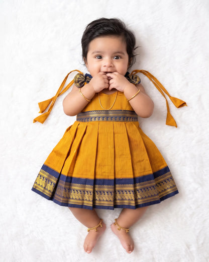 A baby girl lying on a white textured blanket, wearing a mustard yellow pleated traditional dress with navy blue and gold borders and tie details on the shoulders.
