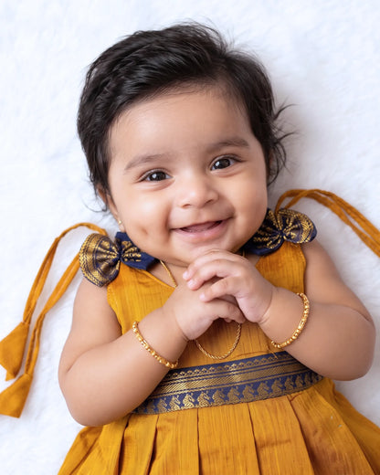 A smiling baby girl lying on a white textured blanket, wearing a mustard yellow dress with a navy blue and gold patterned waistband, bow details on the shoulders, and gold bracelets.