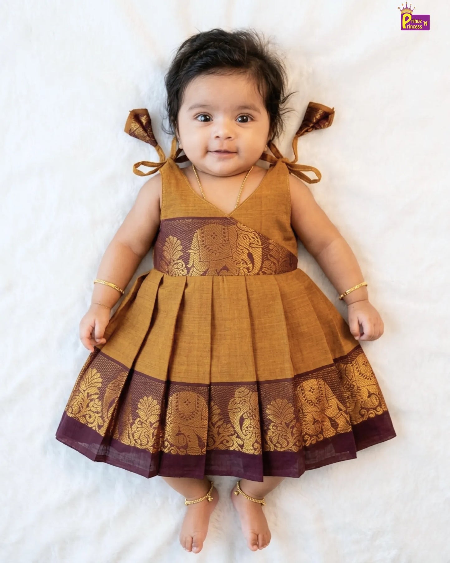 Baby wearing a mustard yellow and maroon traditional dress with elephant patterns on a white background
