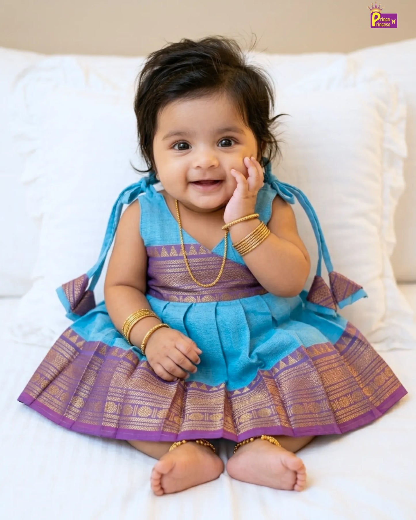 Baby in a blue and purple traditional outfit sitting on a white couch.