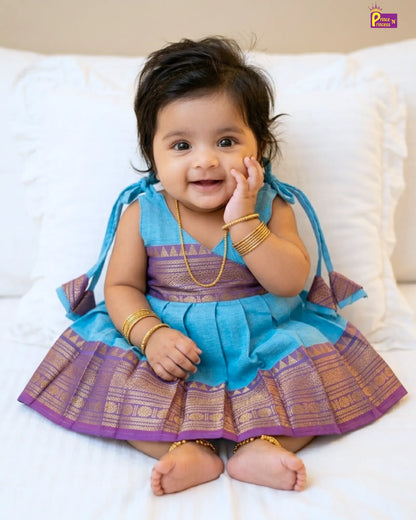 Baby in a blue and purple traditional outfit sitting on a white couch.