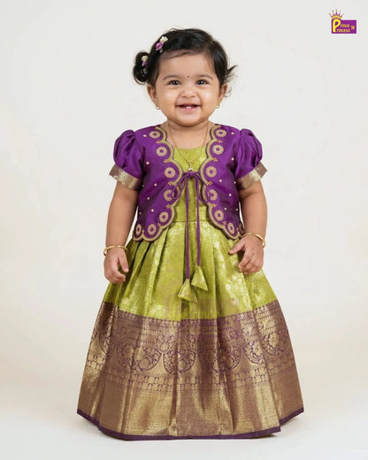 A toddler girl wearing a traditional Indian outfit consisting of a purple embroidered jacket and a lime green and gold skirt, standing against a plain white background.