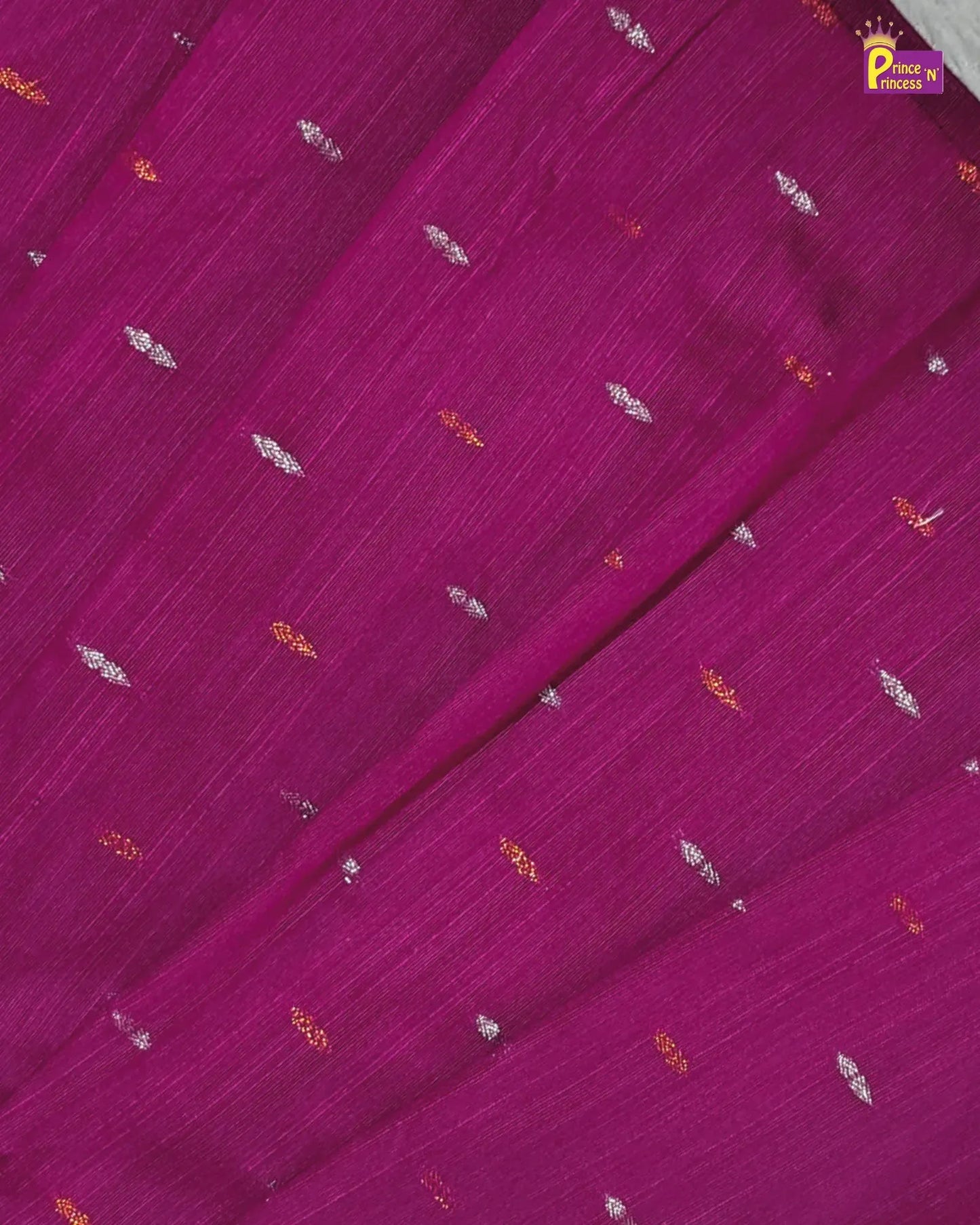 A close-up of a magenta purple fabric with a subtle vertical texture, decorated with small scattered metallic silver and gold zari motifs. A small 'Pook in Delhi' brand logo is visible in the top right corner.
