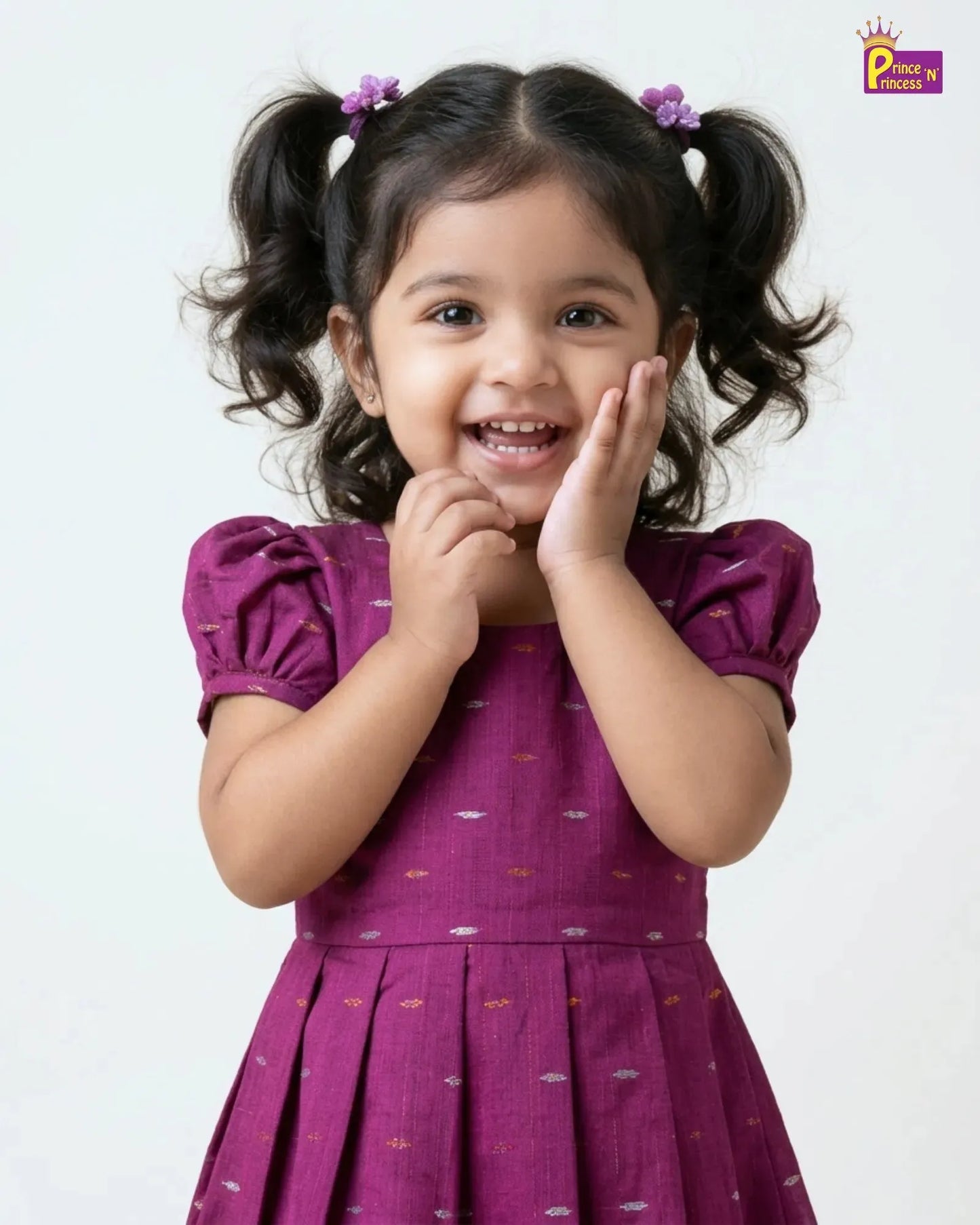 A young girl with dark curly hair in pigtails wearing a purple dress with small colorful cloud prints, posing with her hands near her face against a plain white background.