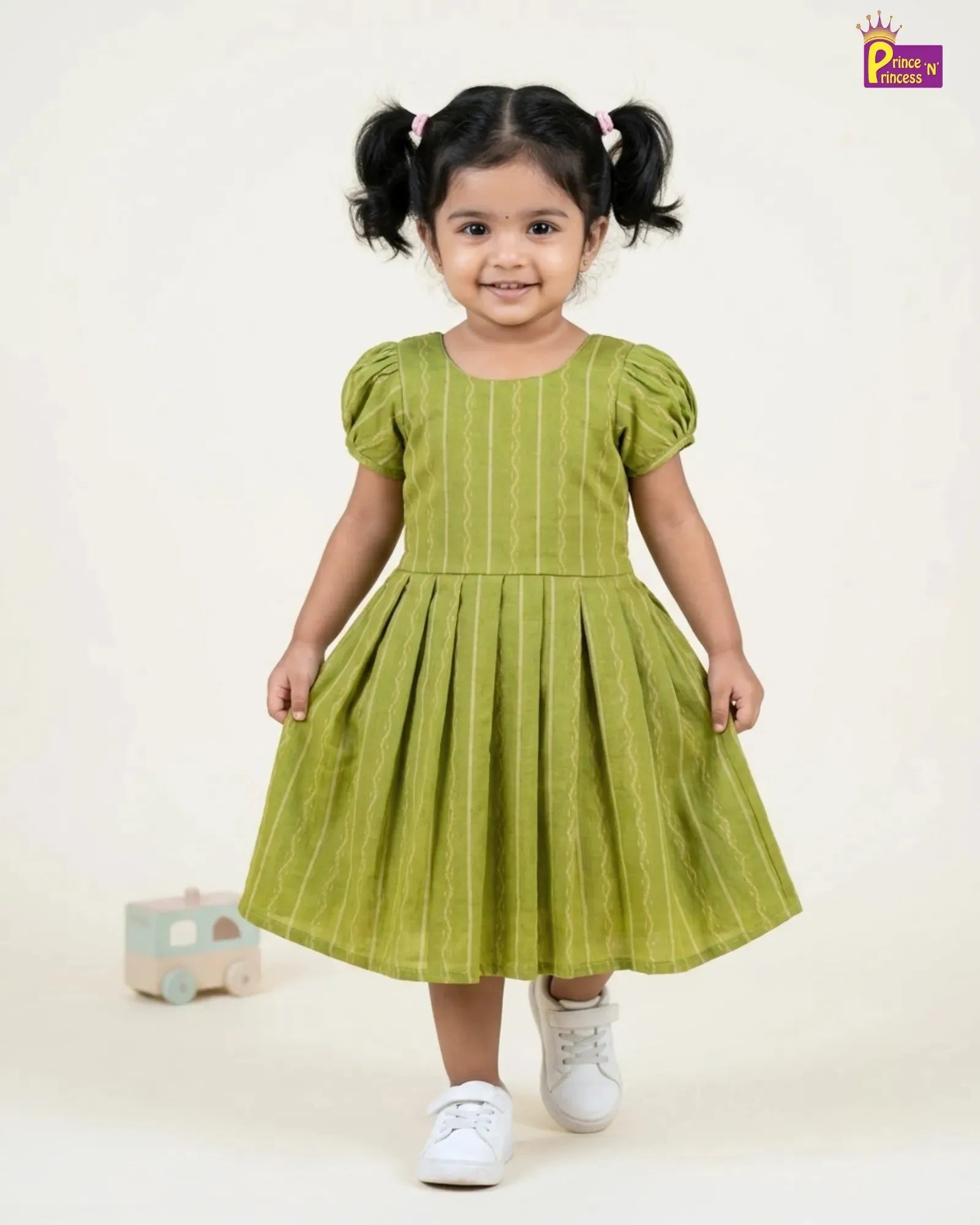 A toddler girl with dark hair in pigtails wearing a light olive green dress with vertical stripes, puff sleeves, and white sneakers, standing against a plain off-white background.