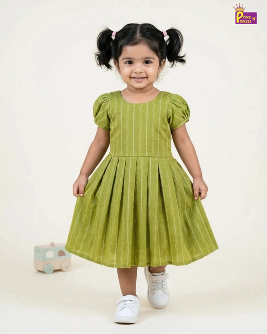 A toddler girl with dark hair in pigtails wearing a light olive green dress with vertical stripes, puff sleeves, and white sneakers, standing against a plain off-white background.