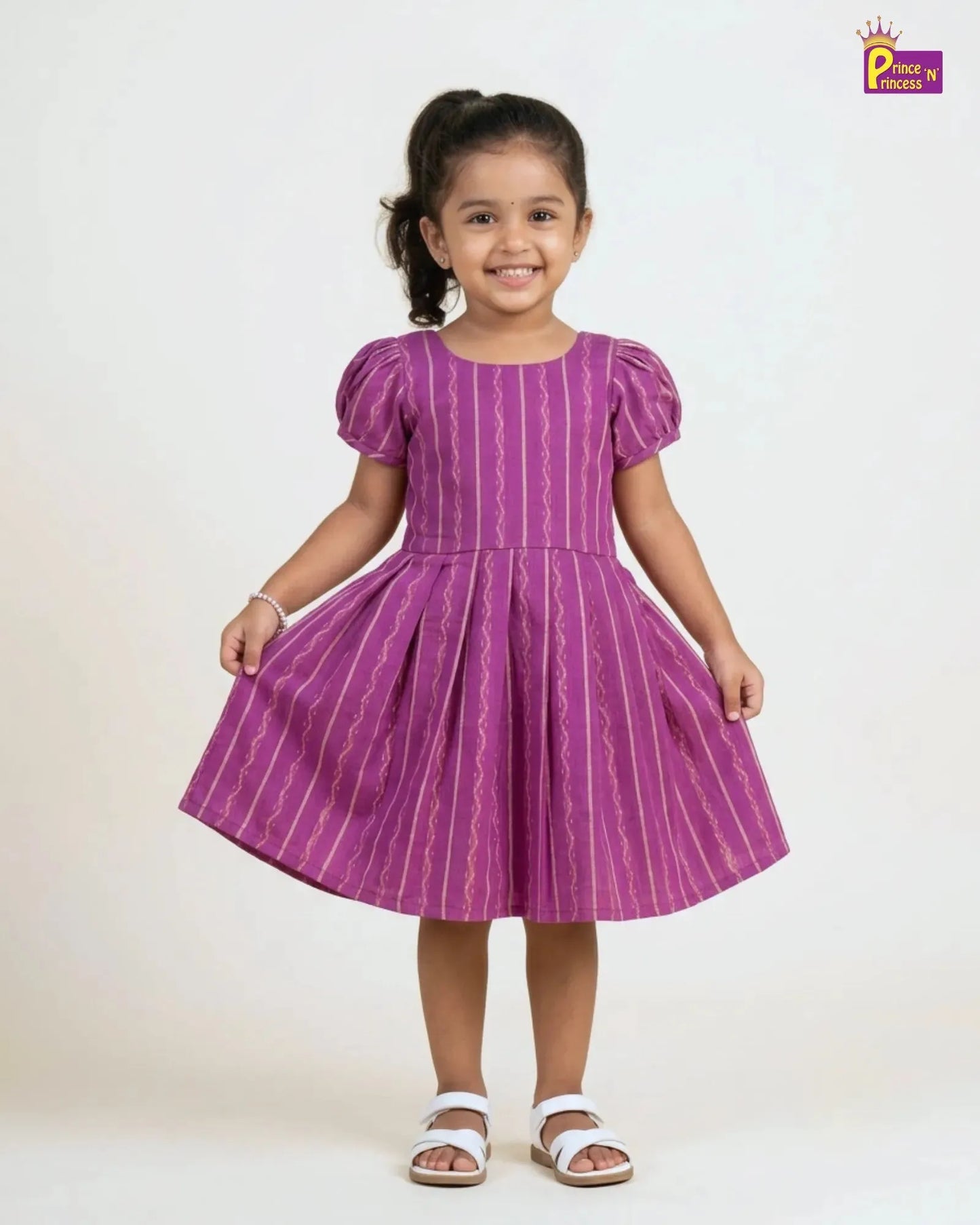 A young girl with her hair in a ponytail stands smiling, wearing a purple short-sleeve dress with vertical white wavy stripes and white sandals, set against a plain light background.