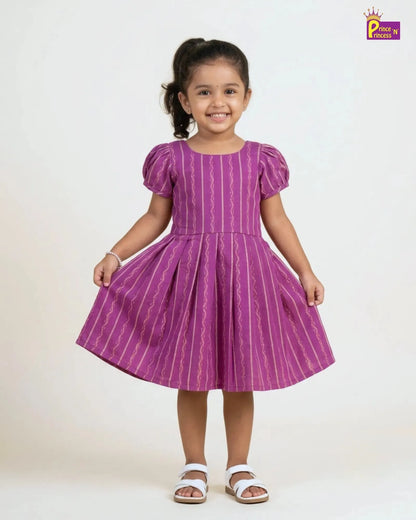 A young girl with her hair in a ponytail stands smiling, wearing a purple short-sleeve dress with vertical white wavy stripes and white sandals, set against a plain light background.