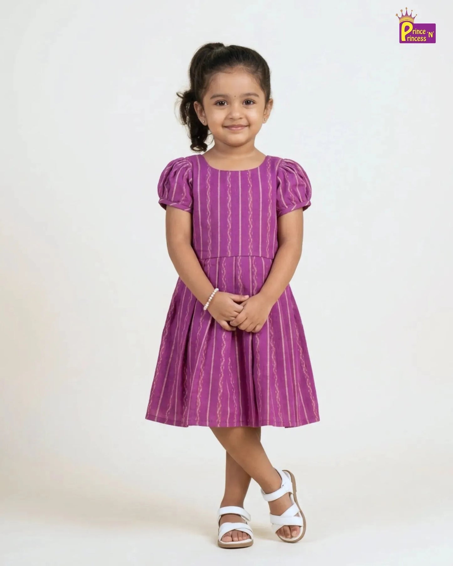 A young girl modeling a purple short-sleeve dress with vertical wavy stripes, standing against a plain white studio background. She is wearing white sandals, and a 'Place by Priya' brand logo is visible in the top right corner.