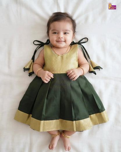 A baby girl lying on a white sheet wearing a sleeveless traditional Indian dress with a metallic gold top, dark green pleated skirt, and black ribbon bows on the back with gold trim.