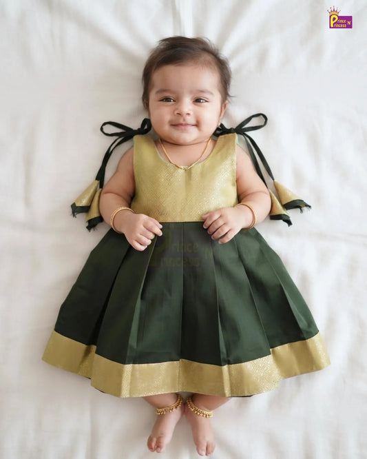 A baby girl lying on a white sheet wearing a sleeveless traditional Indian dress with a metallic gold top, dark green pleated skirt, and black ribbon bows on the back with gold trim.