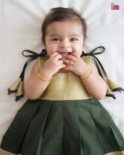 A baby girl lying on a white sheet, wearing a sleeveless green and gold dress with black ribbon bows on the back. She is smiling and touching her mouth, wearing gold bracelets.