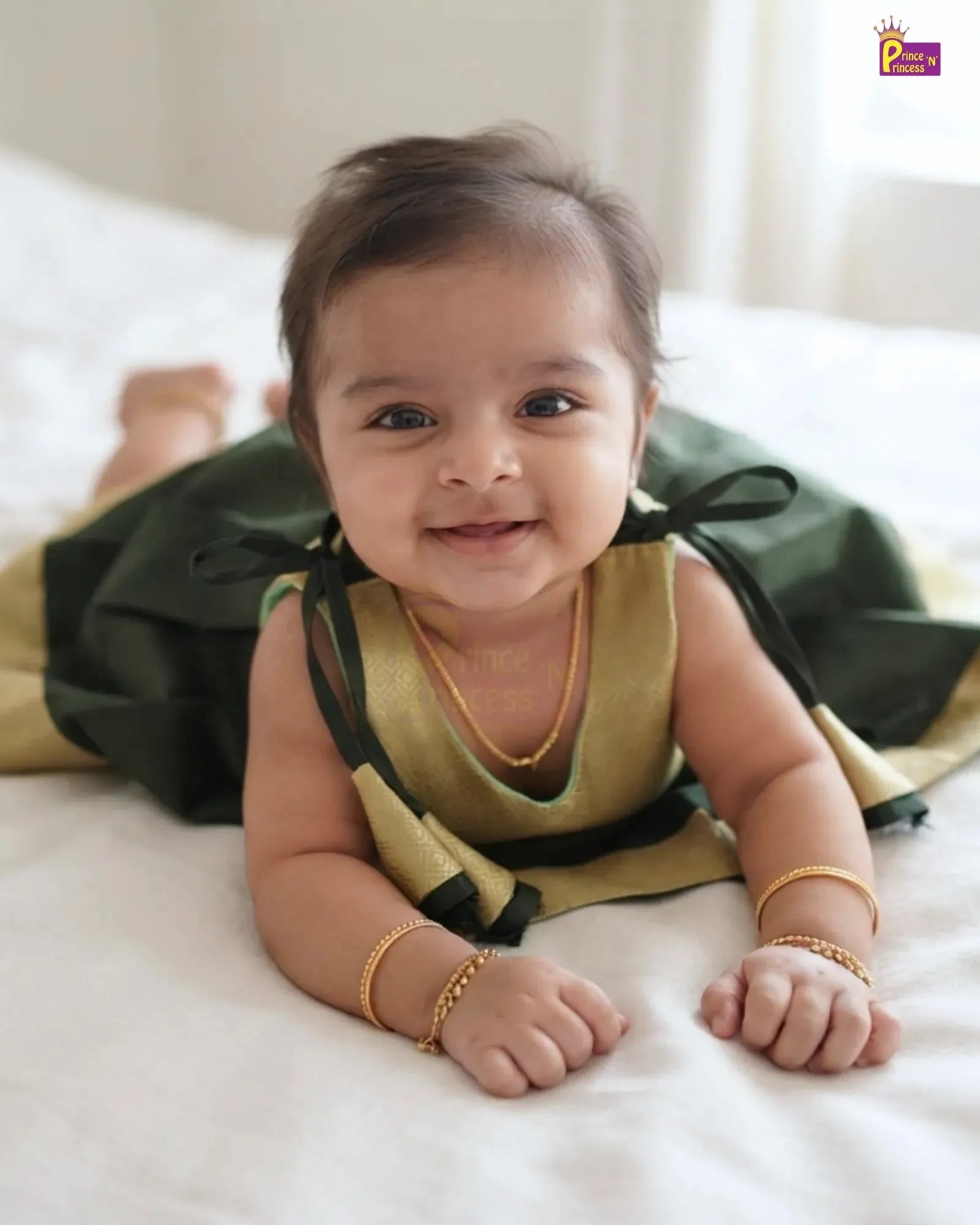 A baby lying on a white bed wearing a gold and dark green traditional outfit with matching gold jewelry.