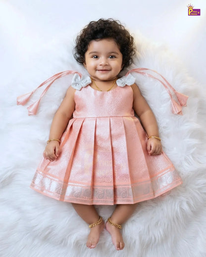 A toddler girl wearing a sleeveless peach-pink pleated dress with white bow details on the shoulders and a silver patterned border on the hem, lying on a white faux fur rug.