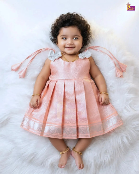 A toddler girl wearing a sleeveless peach-pink pleated dress with white bow details on the shoulders and a silver patterned border on the hem, lying on a white faux fur rug.