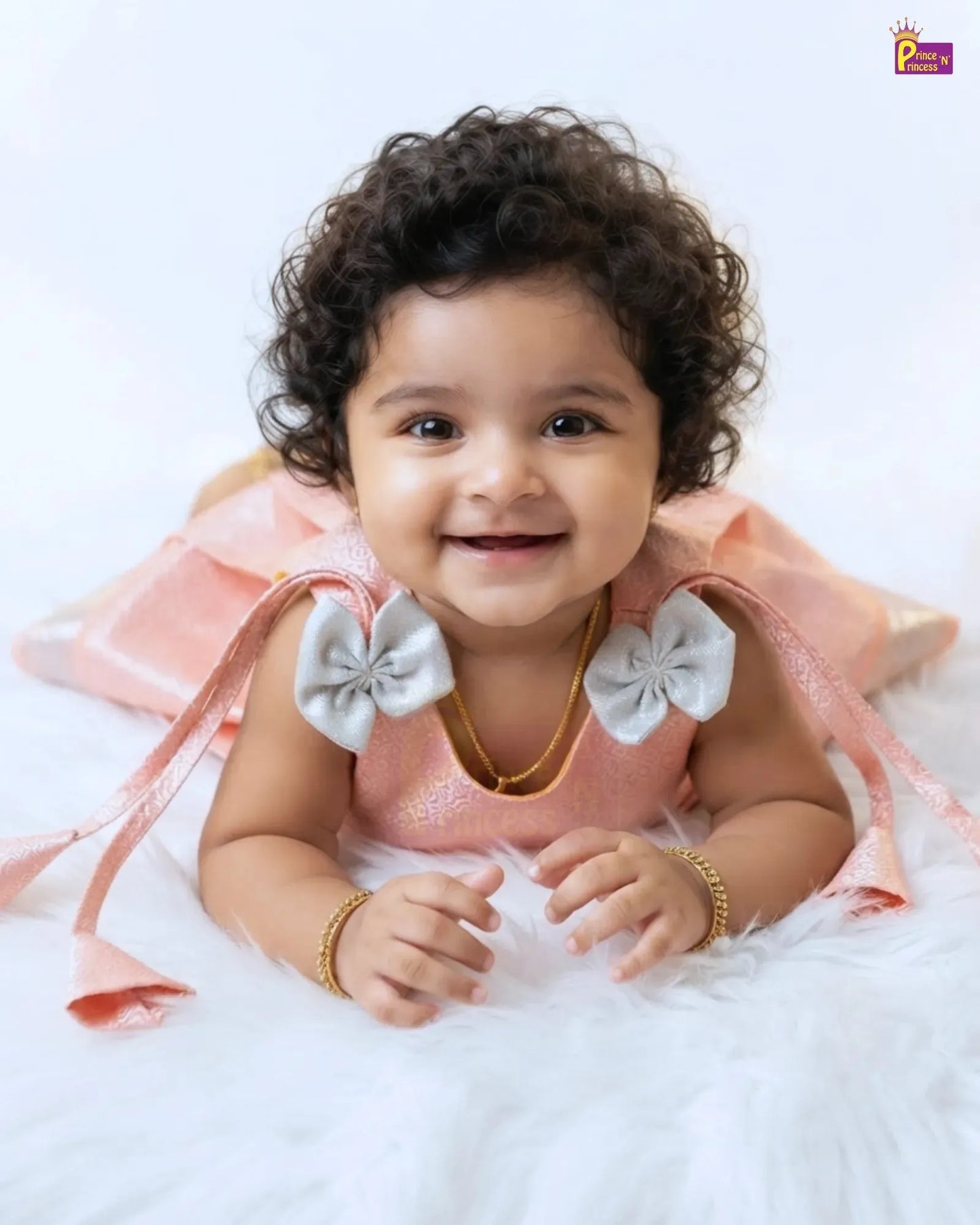 A smiling baby girl with curly dark hair lies on a white fluffy rug wearing a light pink dress with large white bow details on the shoulders and gold jewelry.