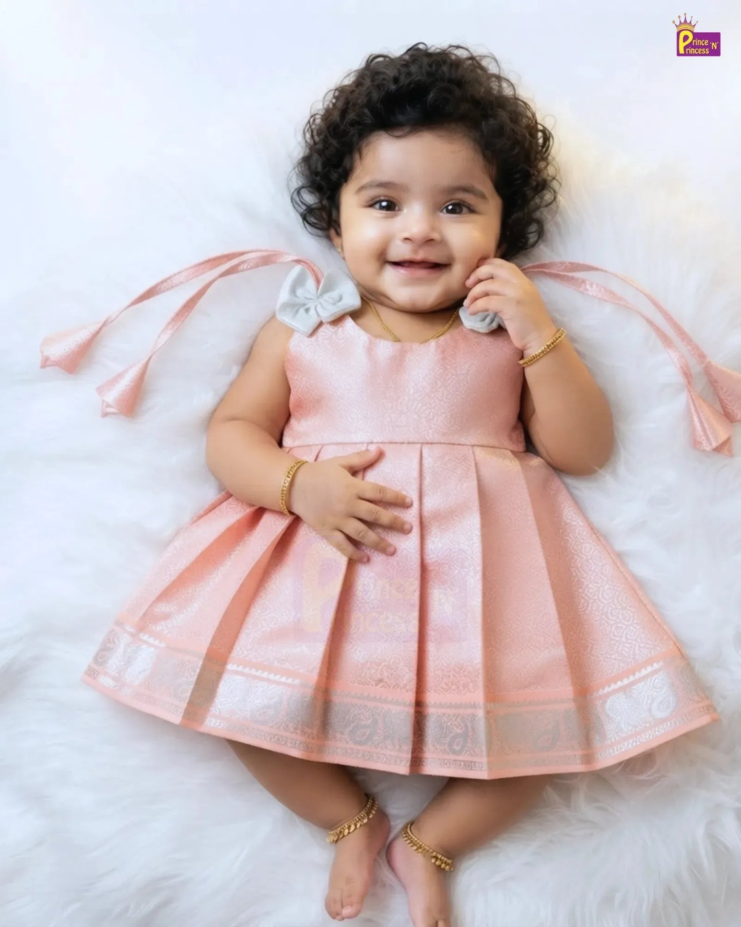 A smiling toddler with curly dark hair lies on a white faux-fur surface wearing a sleeveless peach-pink traditional dress with a pleated skirt, a light gray bow at the neckline, and long ribbon tails. The child is also wearing gold jewelry.