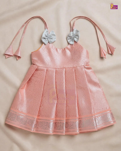 A pink pleated baby dress with a shimmering texture, featuring a decorative silver-gray bow on each strap and a matching patterned hem border, laid flat on a beige fabric background.